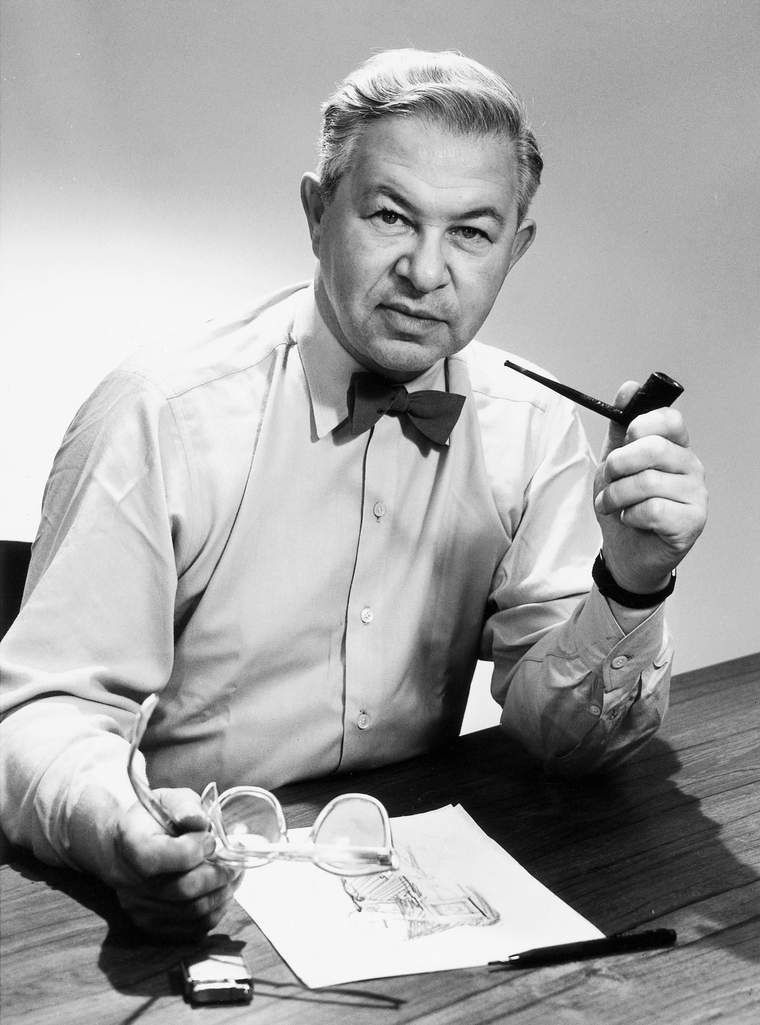 Arne Emil Jacobsen portrait en noir et blanc. C'est un homme aux cheveux courts. Il est assis à un bureau sur lequel se trouve un dessin et un crayon. Il est habillé d'une chemise et d'un noeud papillon. Il tient dans sa main gauche une pipe et dans sa main droite des lunettes. Il a une montre à son poignet gauche.